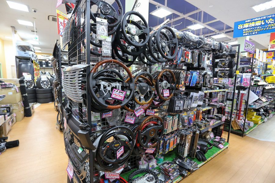 Auto parts store with car steering wheels, accessories, and tires on display. Japanese signs show product information and prices.
