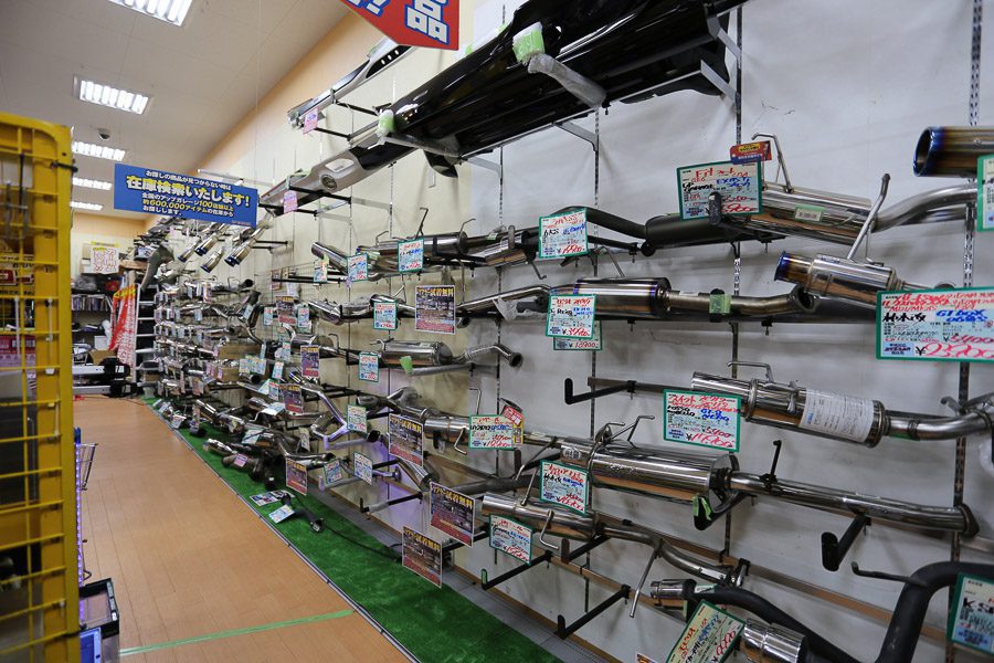 Exhaust pipes and automotive parts displayed on shelves in a Japanese car parts store, with signs listing prices.