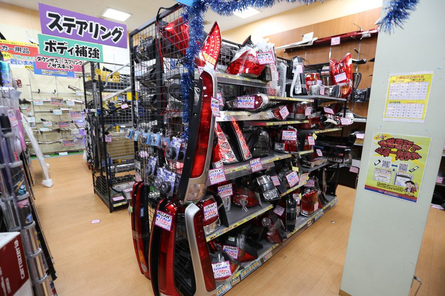 Auto parts store shelf with various car accessories, headlights, and components labeled in Japanese.