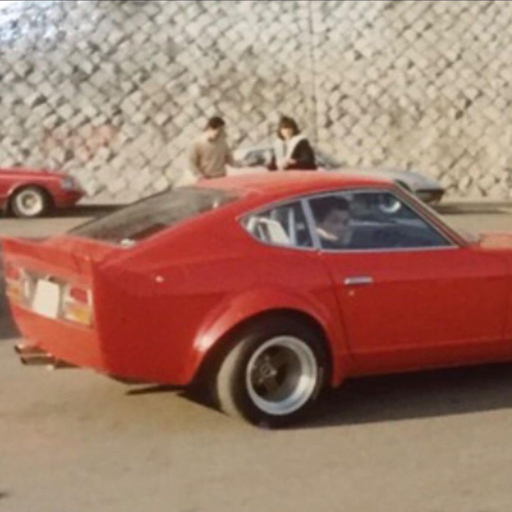Vintage red sports car with flared fenders parked near stone wall, two people in the background, 1970s Japan.