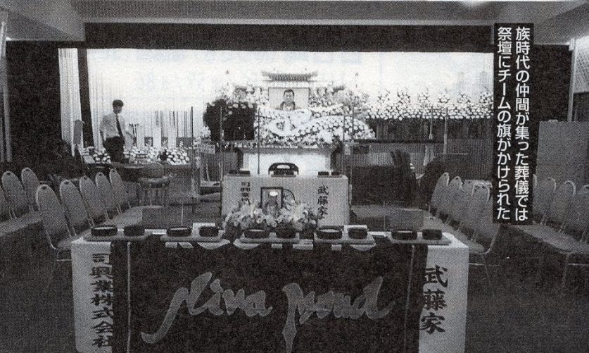 Memorial service with floral tributes and a photo of the deceased, featuring a banner with Japanese text in the background.