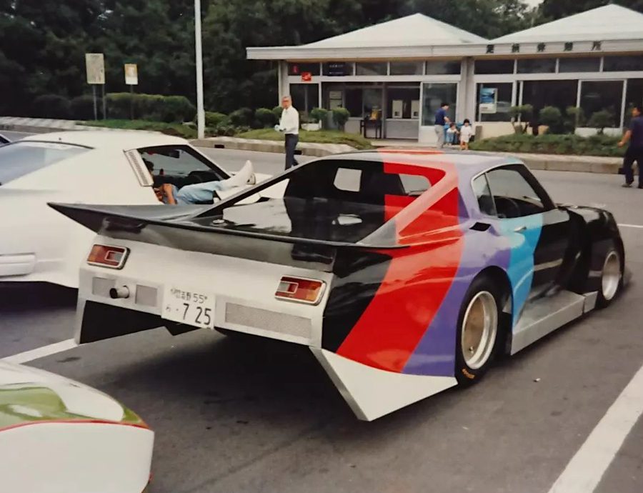 A custom-painted sports car with Japanese plates parked in front of a rest area with a white car nearby.