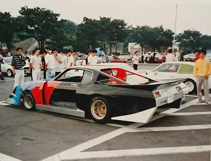 A group of people admires a custom-modified car at a Japanese car meet, featuring vibrant colors and advanced aerodynamics.