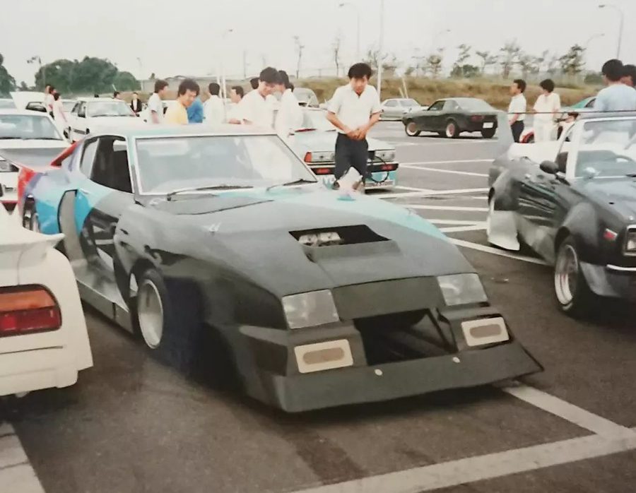 Modified Japanese sports cars at a gathering, with one prominently displayed in the foreground, surrounded by enthusiasts.