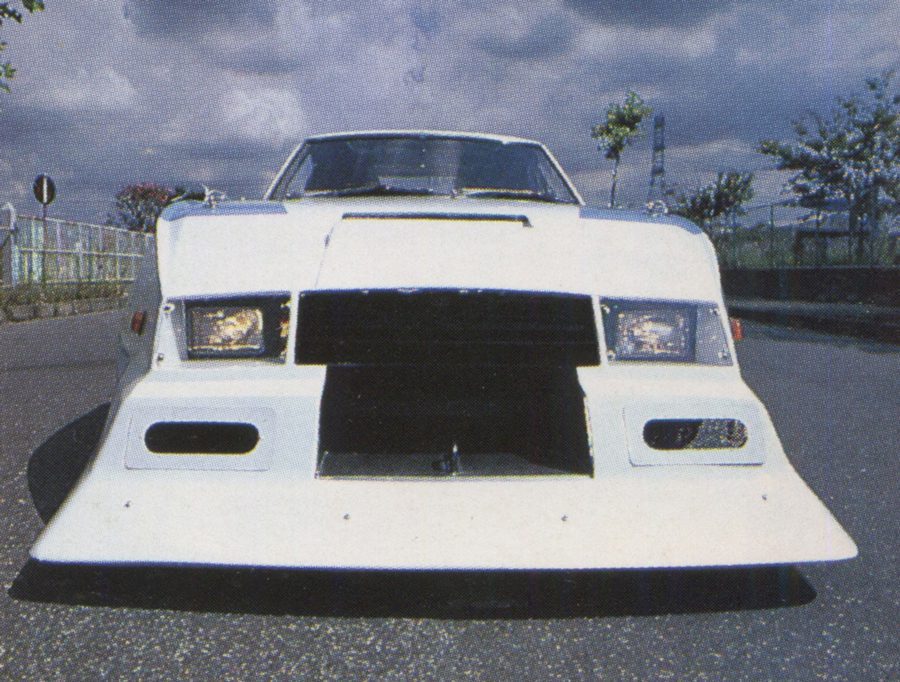 Modified white sports car with aggressive front styling, parked on a street with cloudy skies and trees in the background.