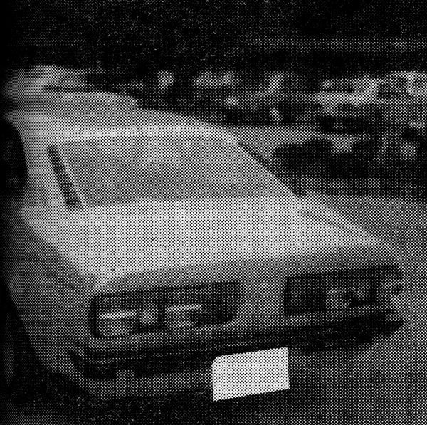 Vintage Japanese car in a grayscale photo with a blurred background of other vehicles in a parking lot.