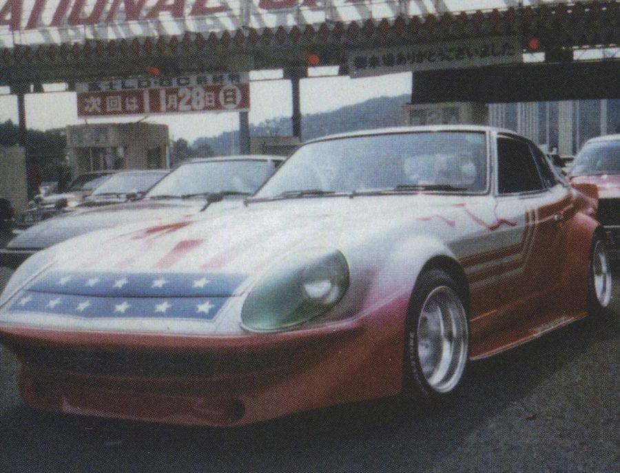 A classic sports car with an American flag paint job in front of a Japanese toll gate at an event.
