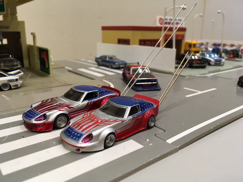 Two American-themed toy cars on a miniature road with buildings and other vehicles in the background.