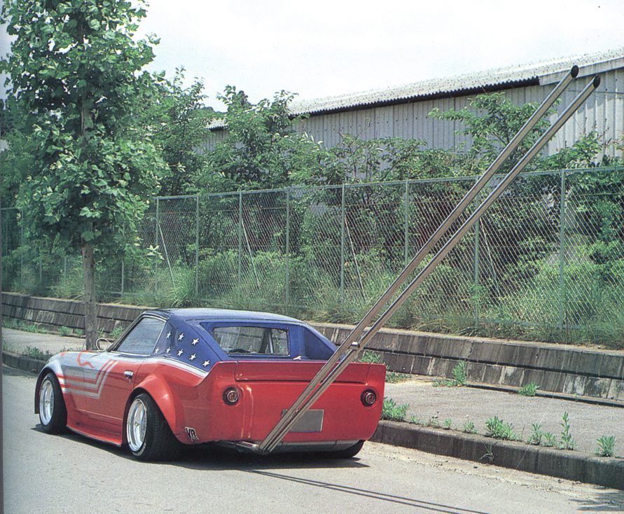 Customized car with extended exhaust pipes, painted in red and blue with star patterns, parked beside a tree and a fence.