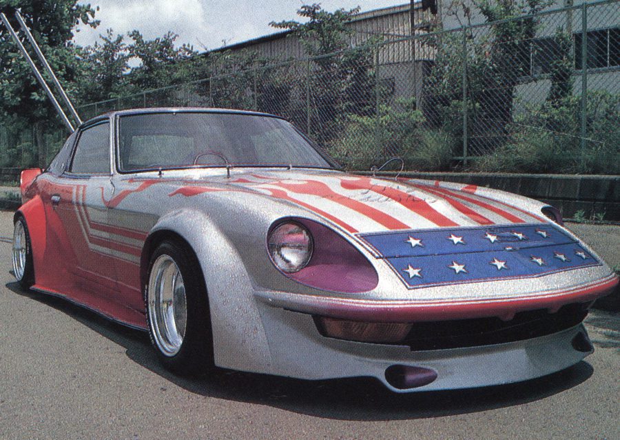 American-themed custom car with flag paint, large rims, and street rods parked on a sidewalk in Japan.