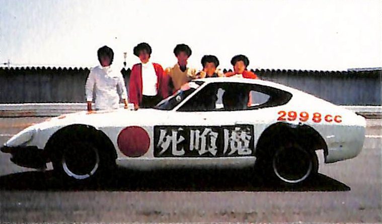 A white sports car with Shinigami and 2998cc written on it, and six people posing behind it on a sunny day.