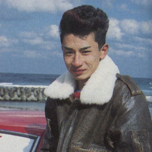 Young man in a shearling leather jacket, standing by a red car near the ocean, with a cloudy sky in the background.
