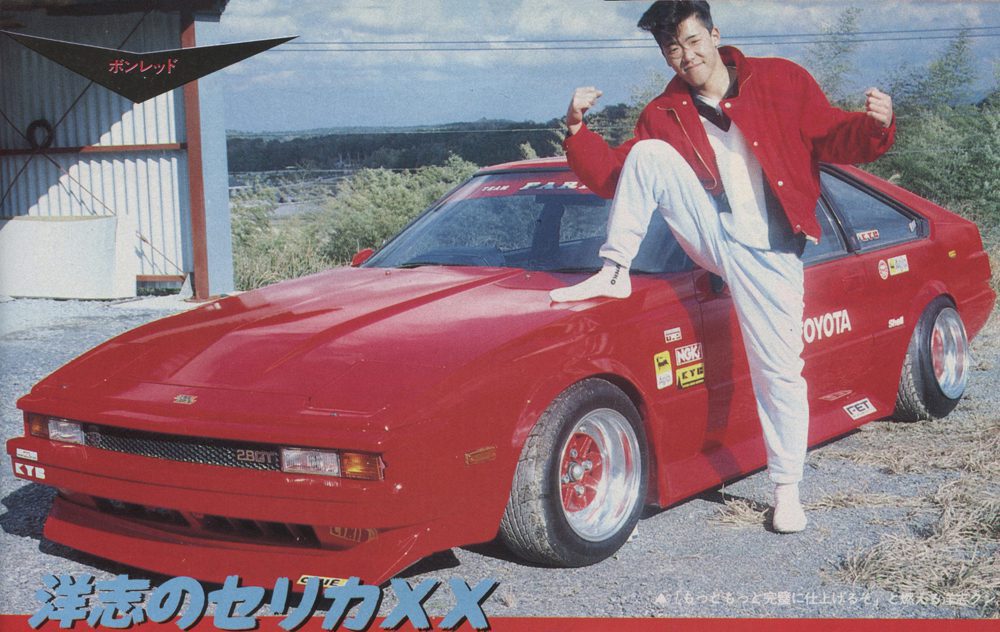 Man in red jacket poses with modified red Toyota Celica XX, showcasing custom decals and parts; Toyota written on the car.