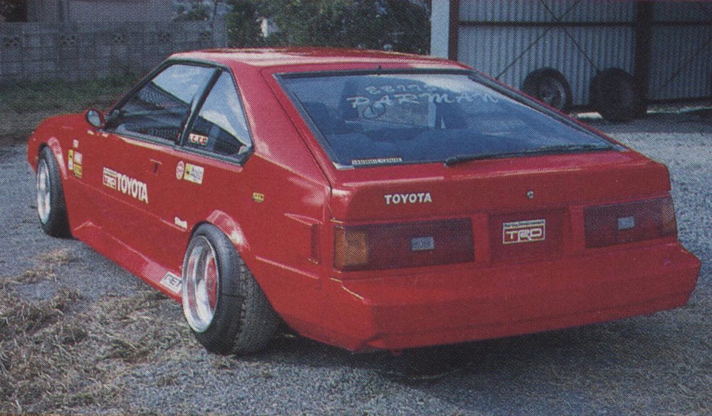 Red Toyota AE86 Trueno with TRD license plate, custom decals, and aftermarket wheels, parked in a gravel lot near a shed.