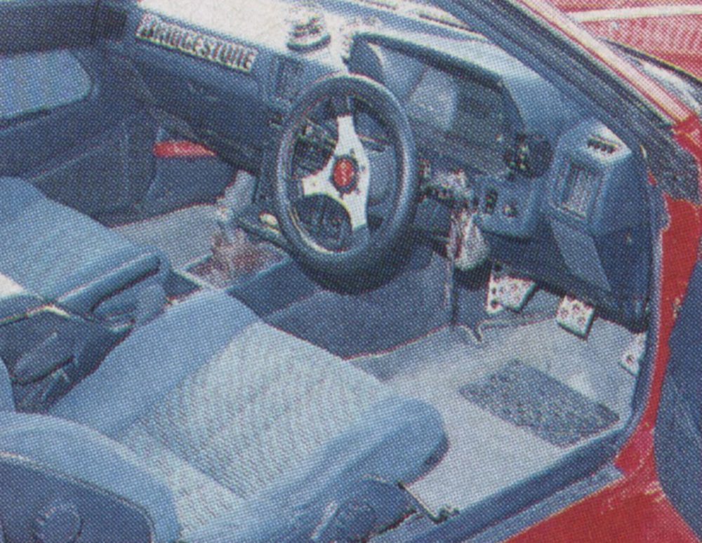 Race car interior with Bridgestone branding, showcasing custom steering wheel, blue seats, and racing pedals.
