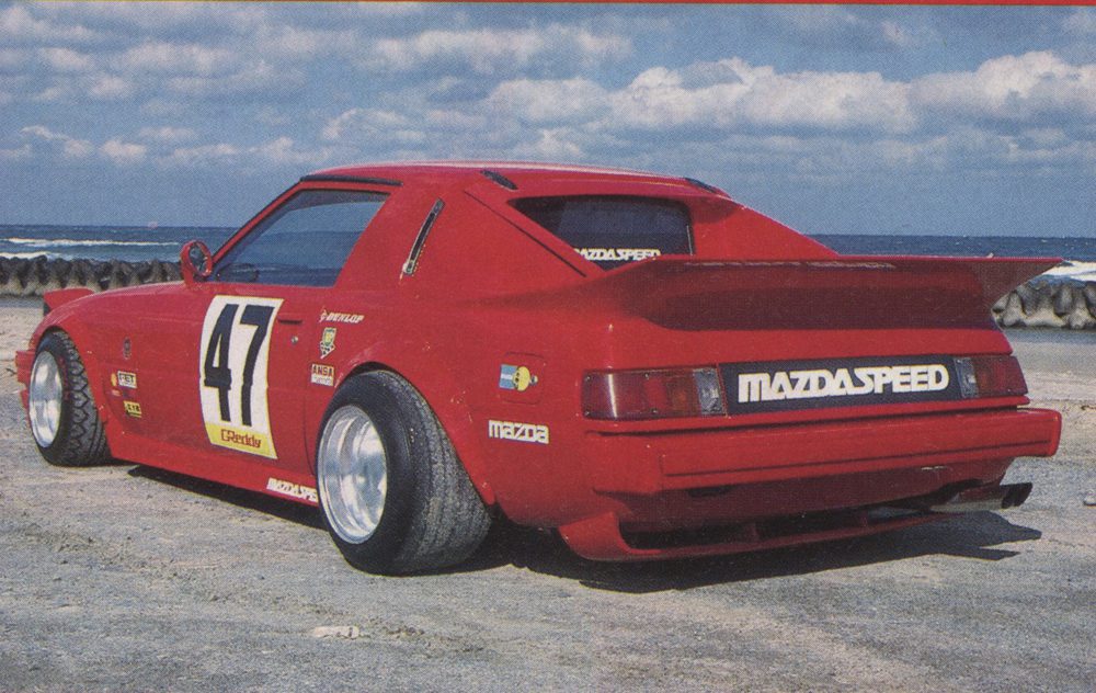 Red Mazda RX-7 with Mazdaspeed decals and number 47, parked near the ocean under a cloudy sky.