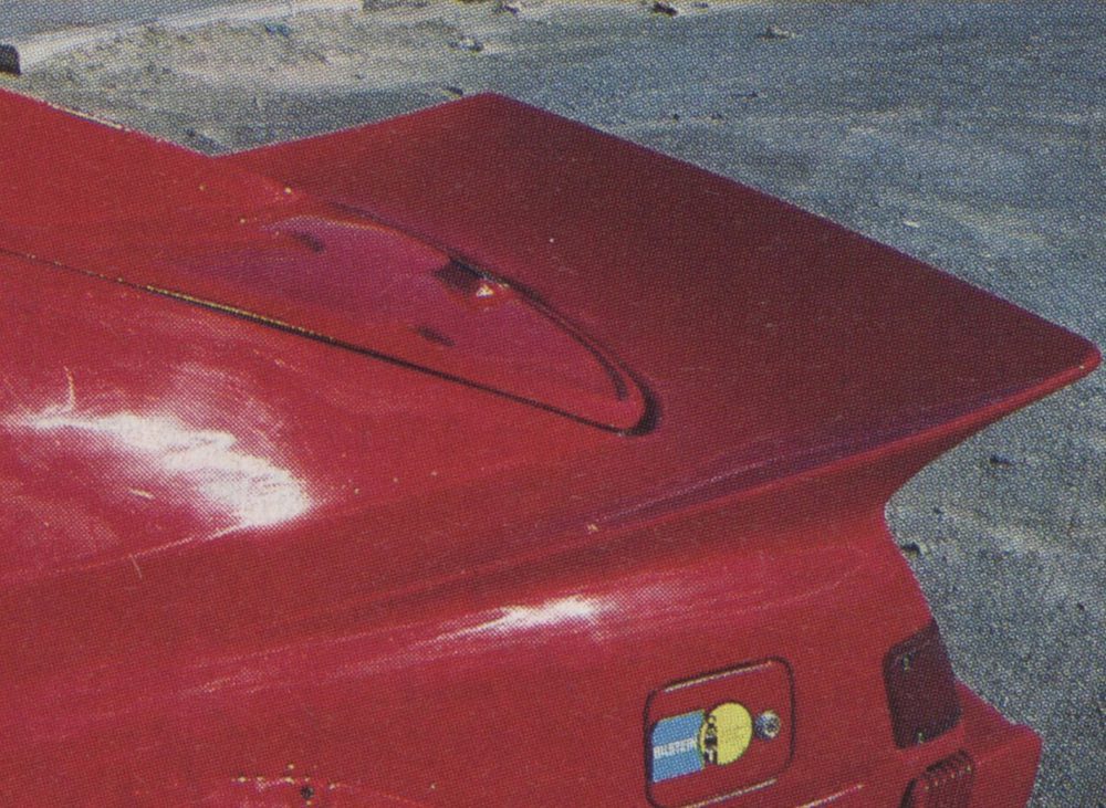 Close-up of the rear spoiler and tail light of a red sports car with Bilstein sticker on a sandy pavement background.