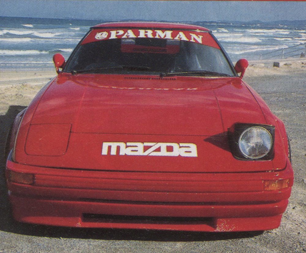 Front view of a red Mazda RX-7 sports car parked by the beach with PARMAN written on the windshield banner.