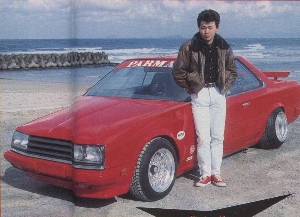 Man standing next to a red Nissan Skyline DR30 Turbo RS with Parma windshield banner on a beach.