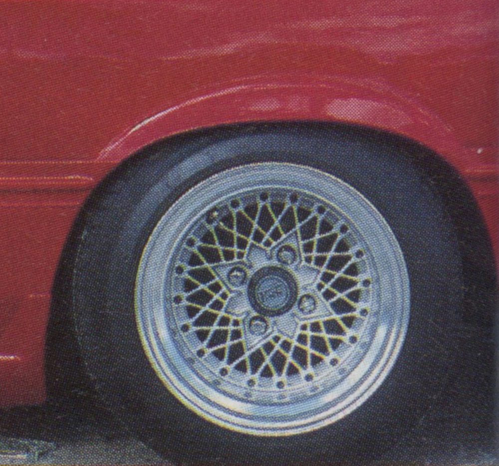 Close-up of a silver alloy wheel with radial spokes on a red car. The center cap has the logo RS Watanabe.