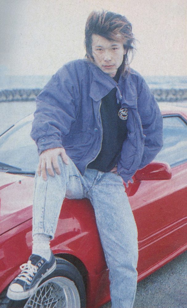 Young person with stylish hair poses on a red sports car, wearing a casual blue jacket and jeans. Fujimi Kaido style.