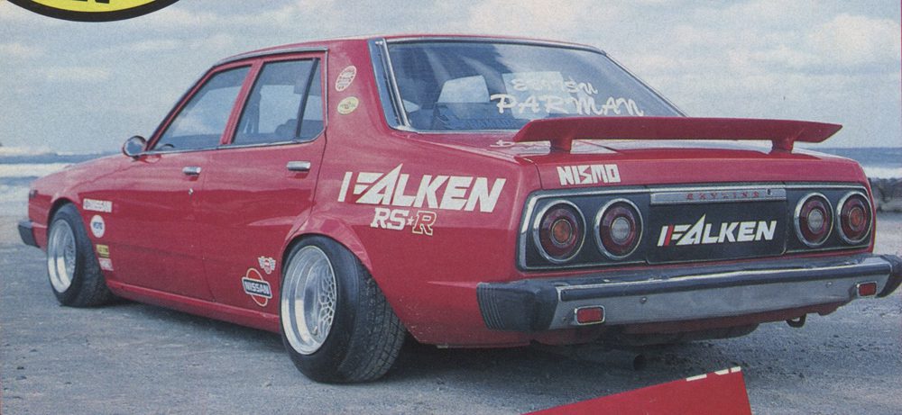 Red modified Nissan Skyline 2000 GT-X with Falken and Nismo decals, rear view, at a beach with ocean in the background.
