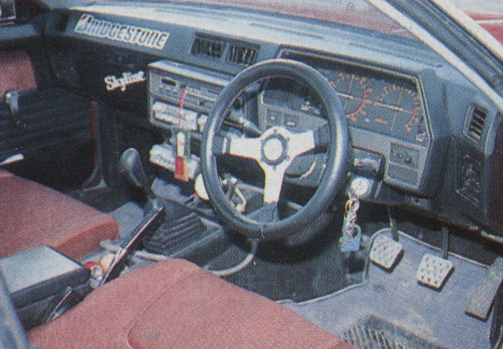 Interior of an old Nissan Skyline featuring a Bridgestone sticker on the dashboard, steering wheel, and pedal assembly visible.