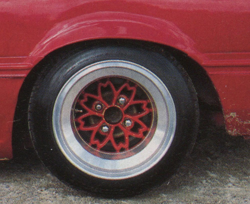 Red car with custom flower-shaped wheel rim design, parked on a concrete surface. Unique automotive detailing.