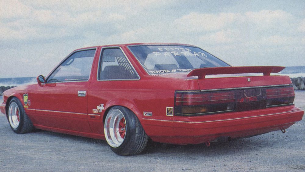 Red Toyota classic sports car with custom modifications near ocean, featuring ミスターR (Mr. R) and パーマン (Parman) decals.