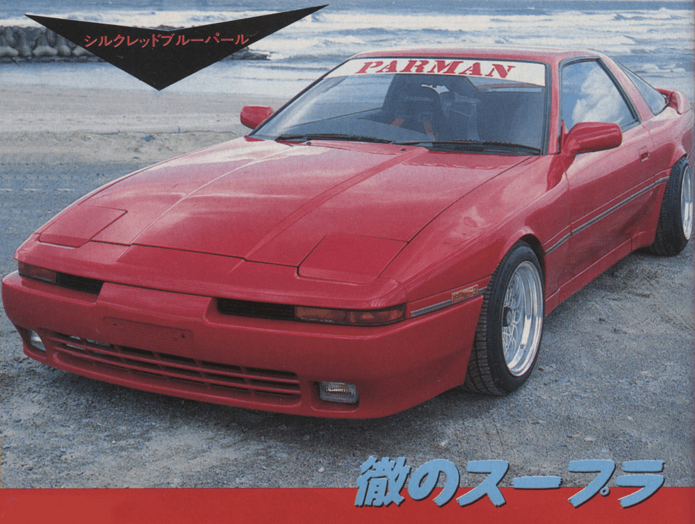 Red Toyota Supra parked by the beach, Silk Red Blue Pearl and Parman on windshield. Japanese text means Supra's Pride.
