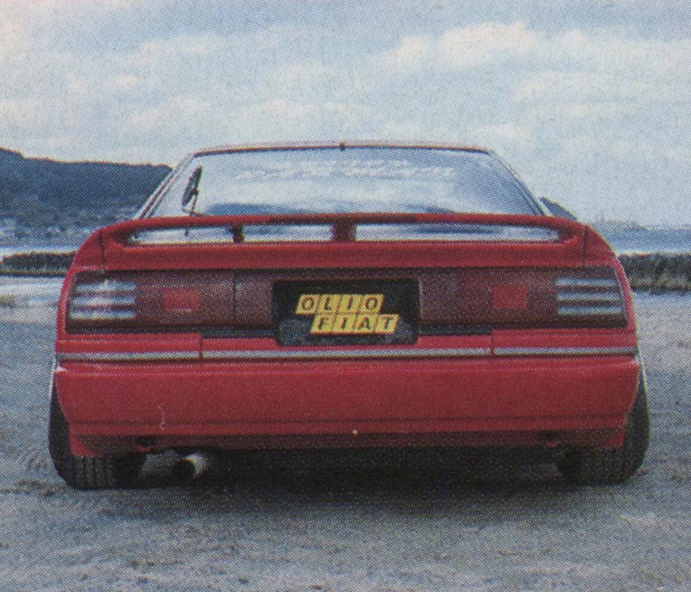 Rear view of a red sports car with OLIO FIAT on the license plate, parked by the beach with mountains in the background.