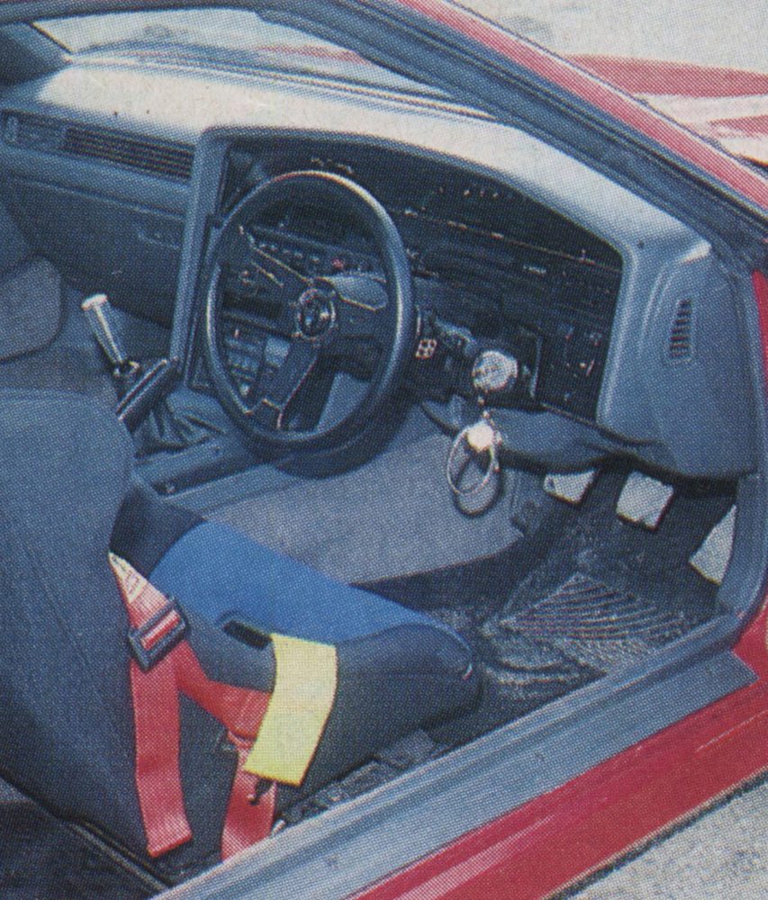 Interior dashboard and steering wheel of a 1980s sports car, showcasing gear shift and a colorful seatbelt harness.