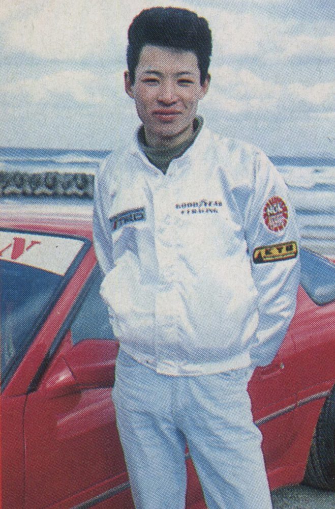 Young man in a Goodyear jacket posing by a red sports car at the beach. Text on jacket: GOODYEAR and TRD (Toyota Racing Development).