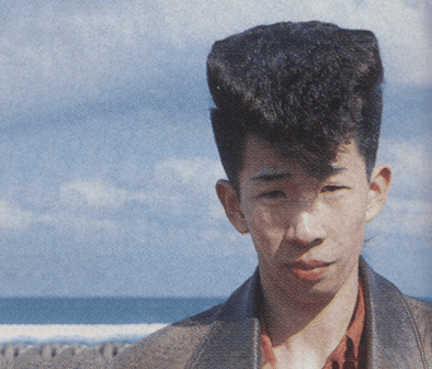 Young man with unique tall hairstyle standing outdoors with ocean and cloudy sky background.