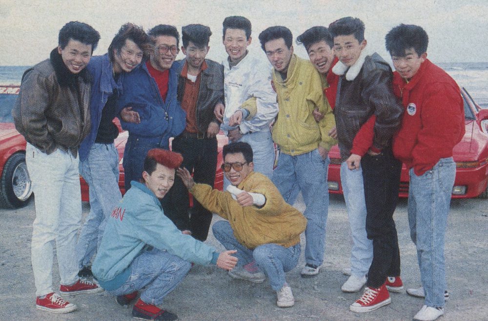 Group of friends with retro fashion at a beach, posing near red sports cars. Japan text seen on a blue jacket.