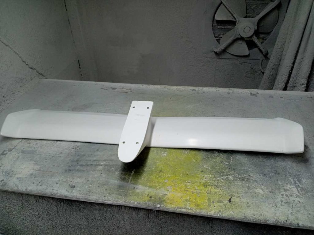 Freshly painted white car spoiler in a workshop. Industrial fan and concrete walls in the background.