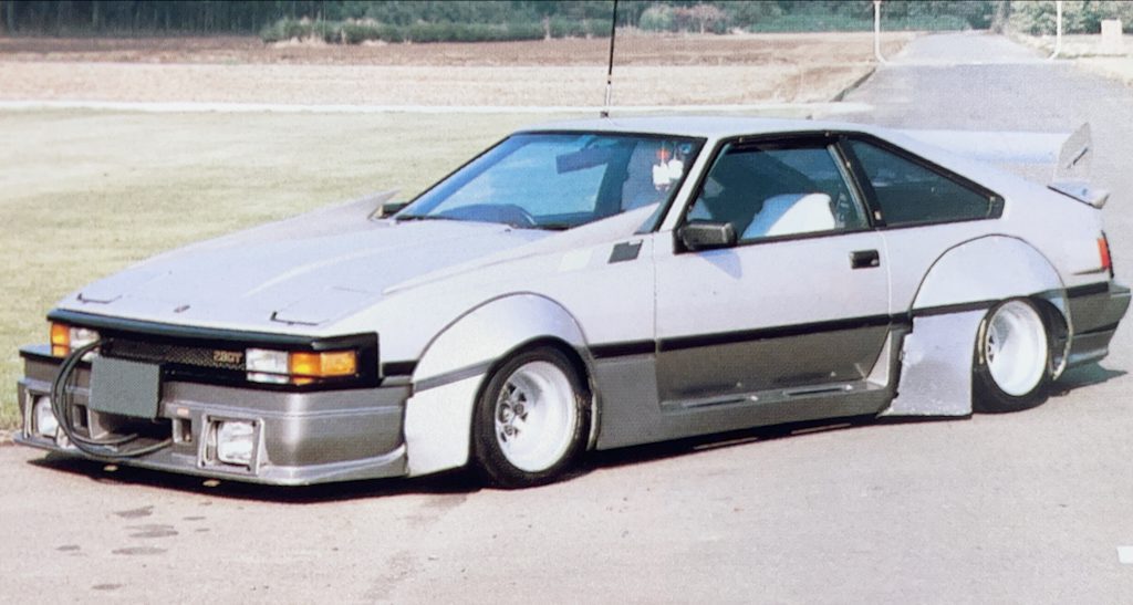 Customized retro Japanese Toyota Celica XX car with wide  Savanna works fenders and a low stance parked on a rural road with green fields in the background.