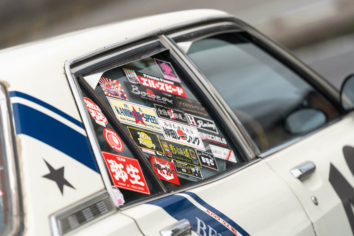 A white car window with various colorful stickers, including Japanese text and designs like スピード (Speed) and ミッティ (Mitty).