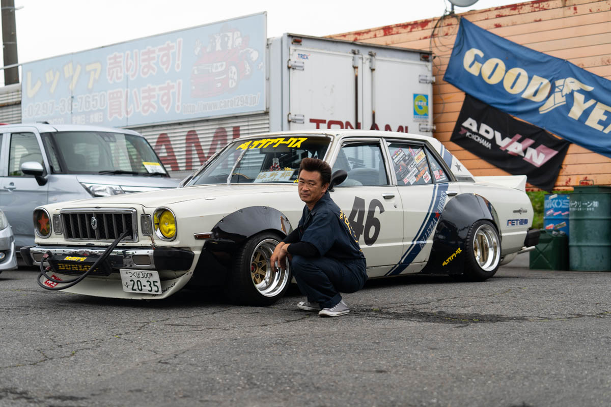 Man kneeling beside a customized vintage car with racing stickers and the number 46, parked near Goodyear and Toyota signs.