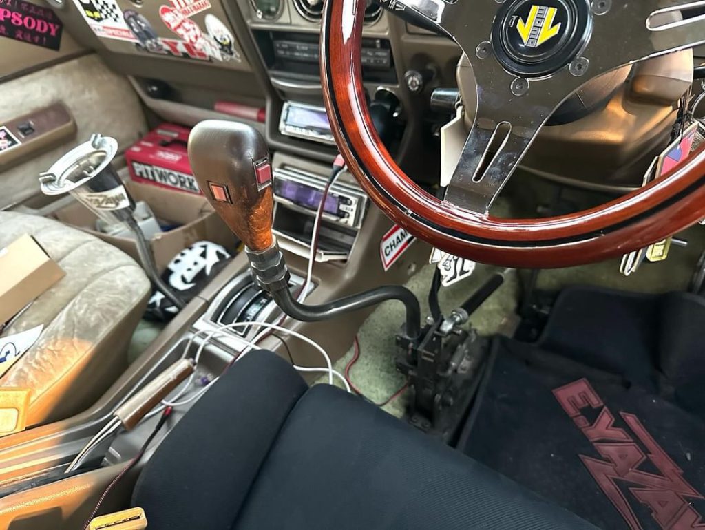 Custom car interior with retro steering wheel, wood shift knob, and dashboard stickers, featuring Champion and Pit Work labels.