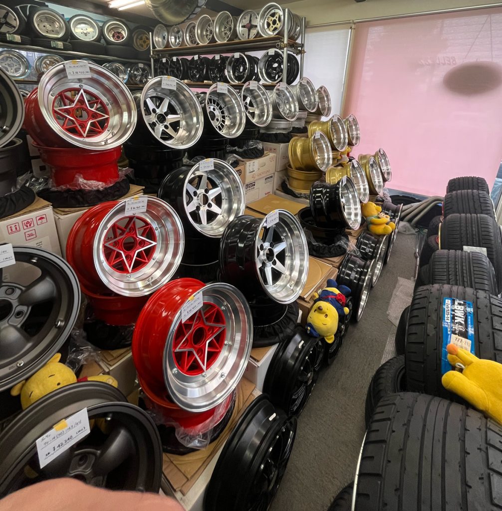 A variety of colorful car rims on display in a Japanese auto parts store, with stacks of tires visible in the foreground.