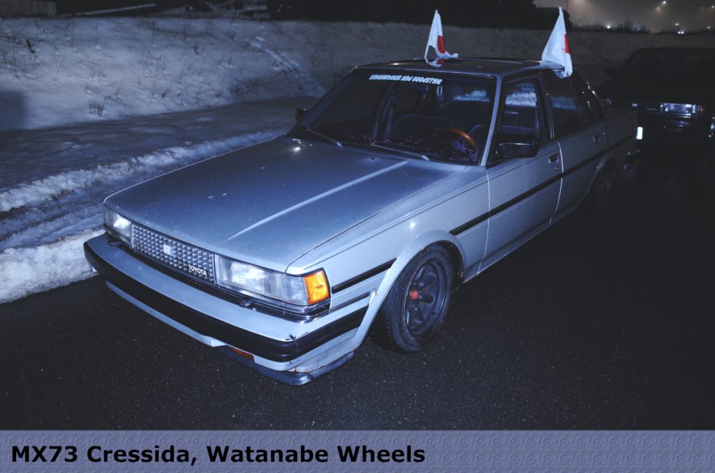 Vintage MX73 Cressida with Watanabe wheels, decked with Japanese flags, and どう見てもアレ対策 on the windshield.