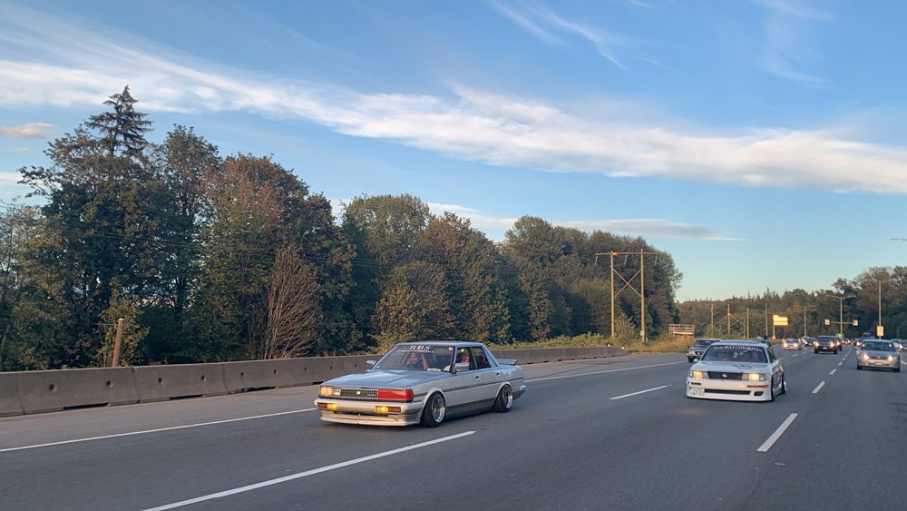Two modified cars driving on a highway during sunset, surrounded by trees and light traffic.