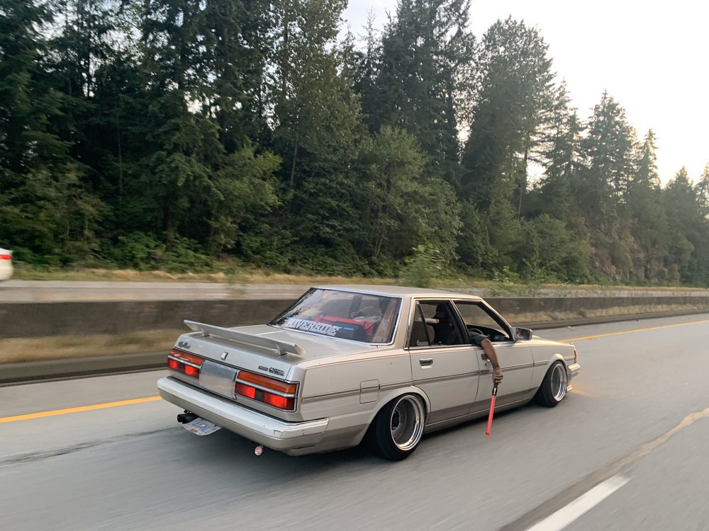 Retro sedan cruising down a highway with a scenic forest backdrop and a person holding a red object out the window.