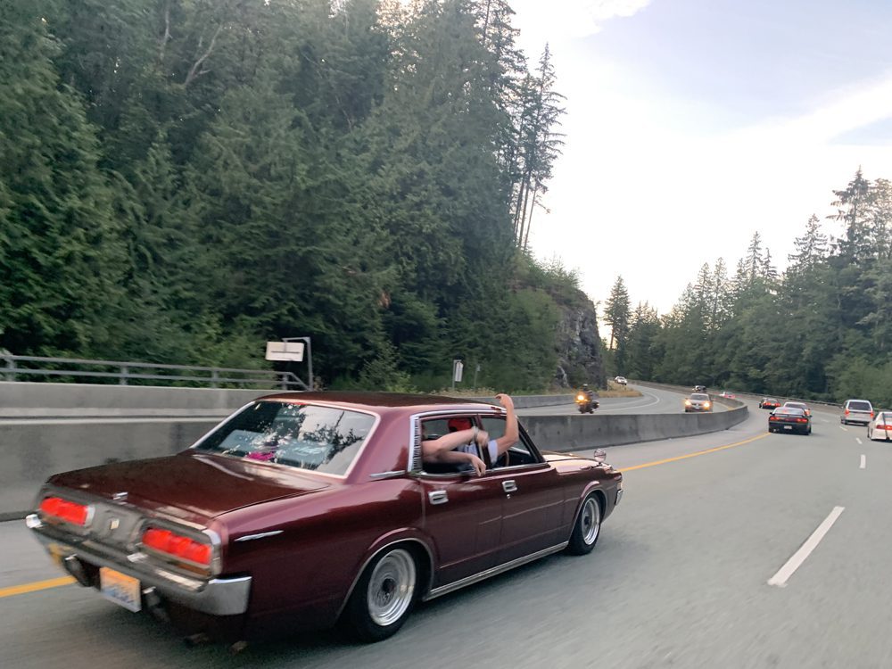 Red vintage car cruising a scenic forest highway, arm out the window, with cars ahead.