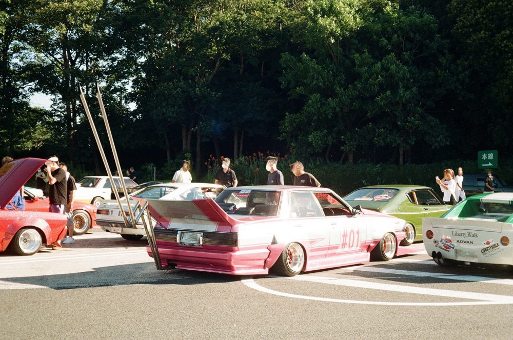 Custom Japanese cars with distinctive paint jobs at an outdoor auto meet, surrounded by people.