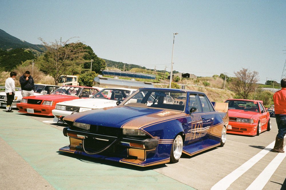 Modified cars parked at a show, featuring a blue and gold Toyota with custom bodywork.