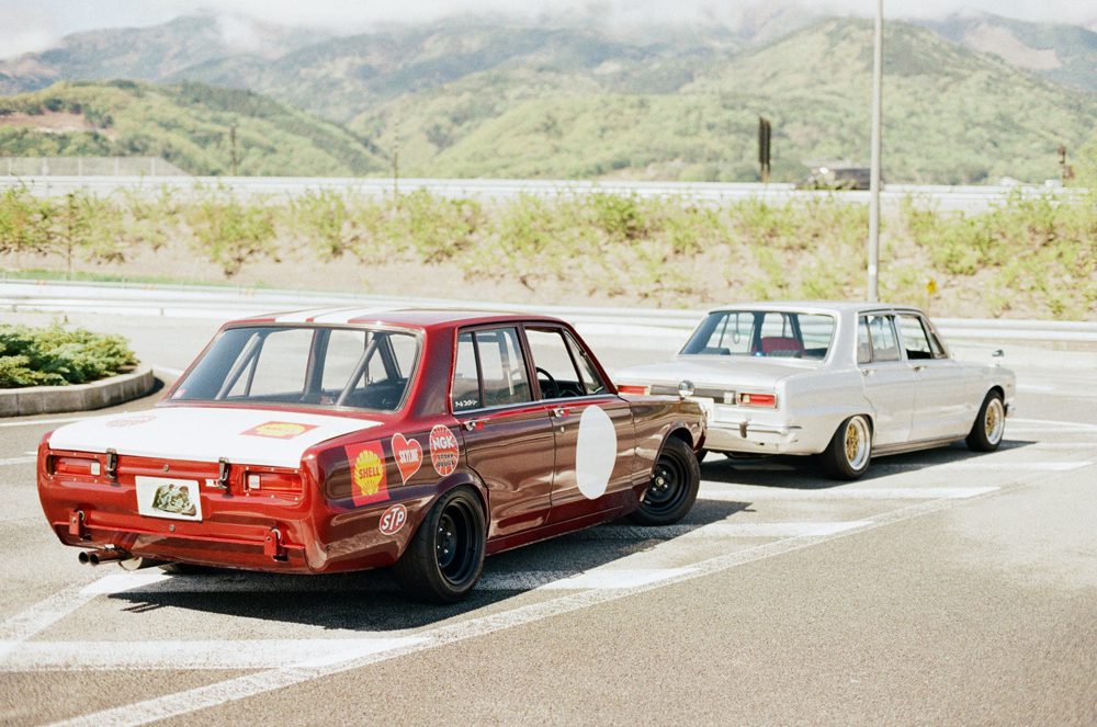 Vintage race cars parked on a scenic road with mountains in the background, showcasing classic automotive style.