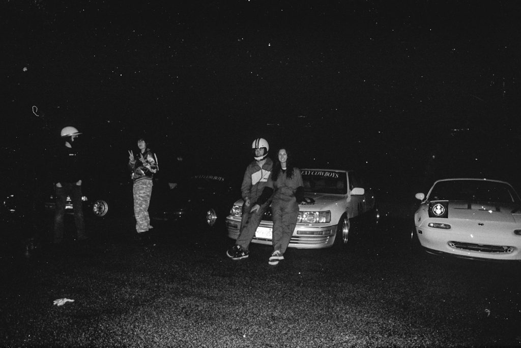 A group of people with modified cars at night, with ATTACK COWBOYS written on windshields.