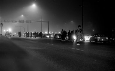 A late-night gathering of people and cars under streetlights in a foggy environment, with signs and 20km speed limit visible.